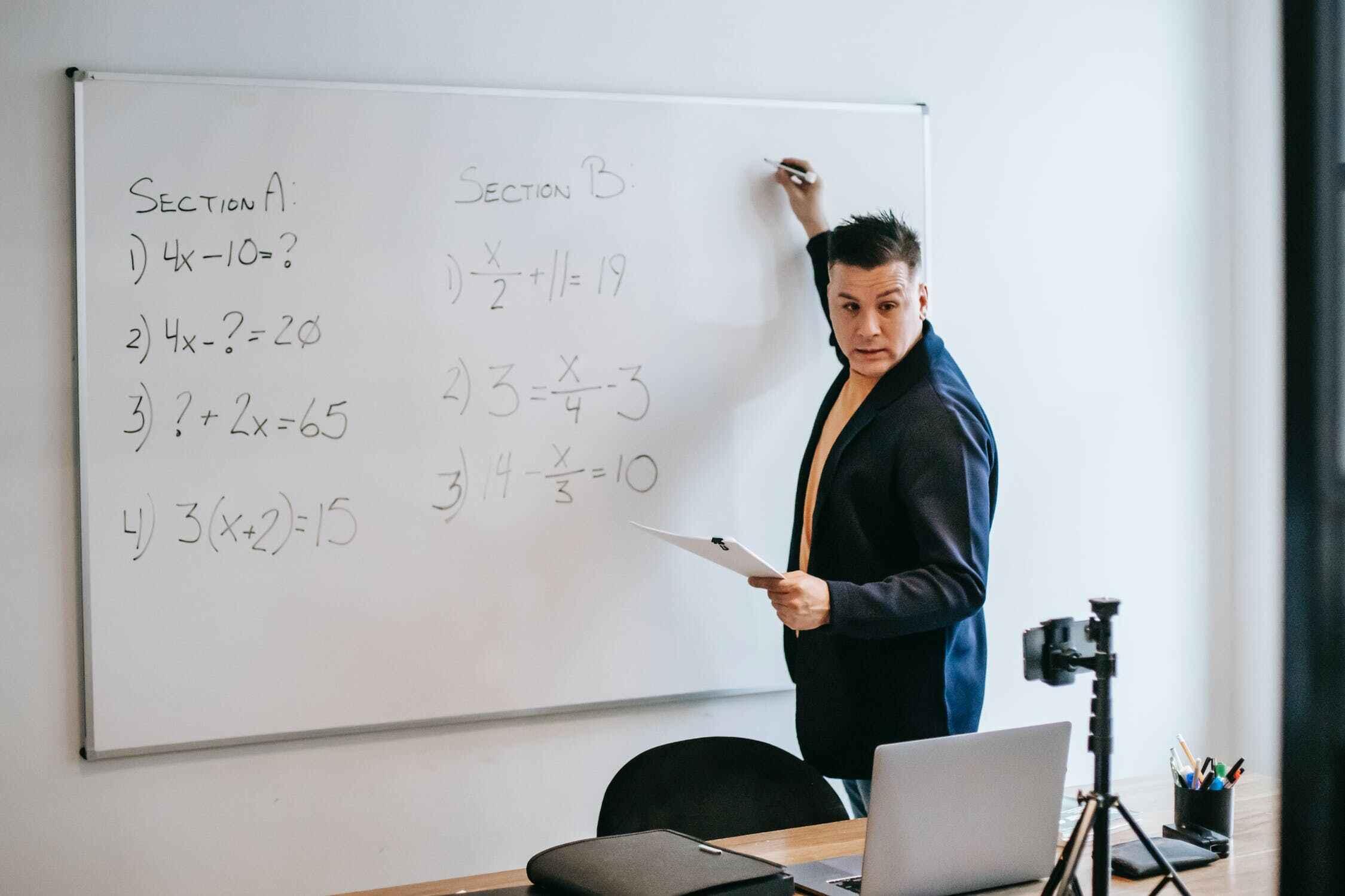 An A level Maths tutor solves a calculation on a whiteboard while delivering an online tutoring lesson.