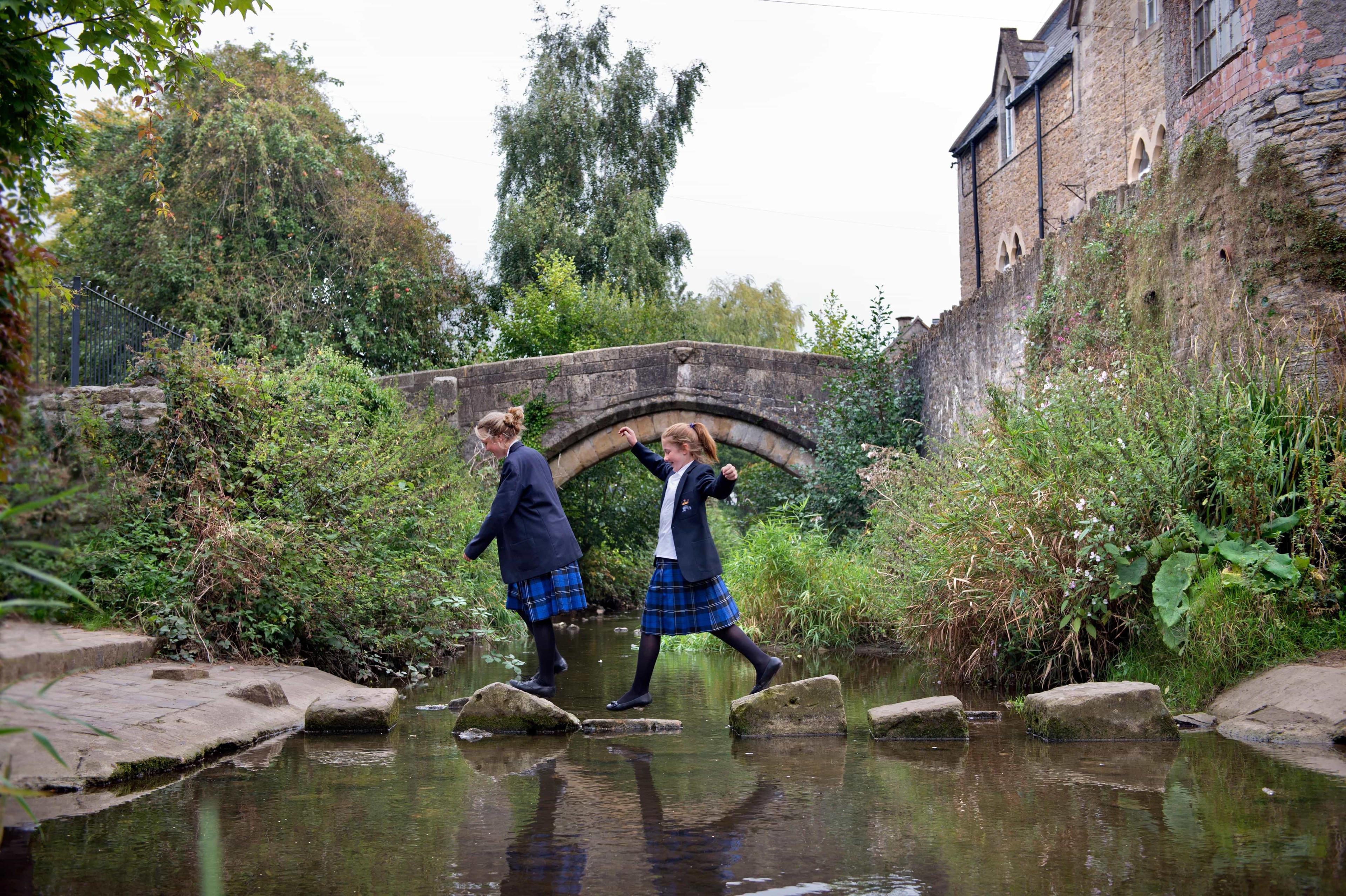 Prep school students jump across stones to cross a stream that runs through the grounds of their school.